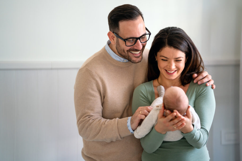 Portrait of young happy man and woman holding newborn cute babe dressed in white unisex clothing.
